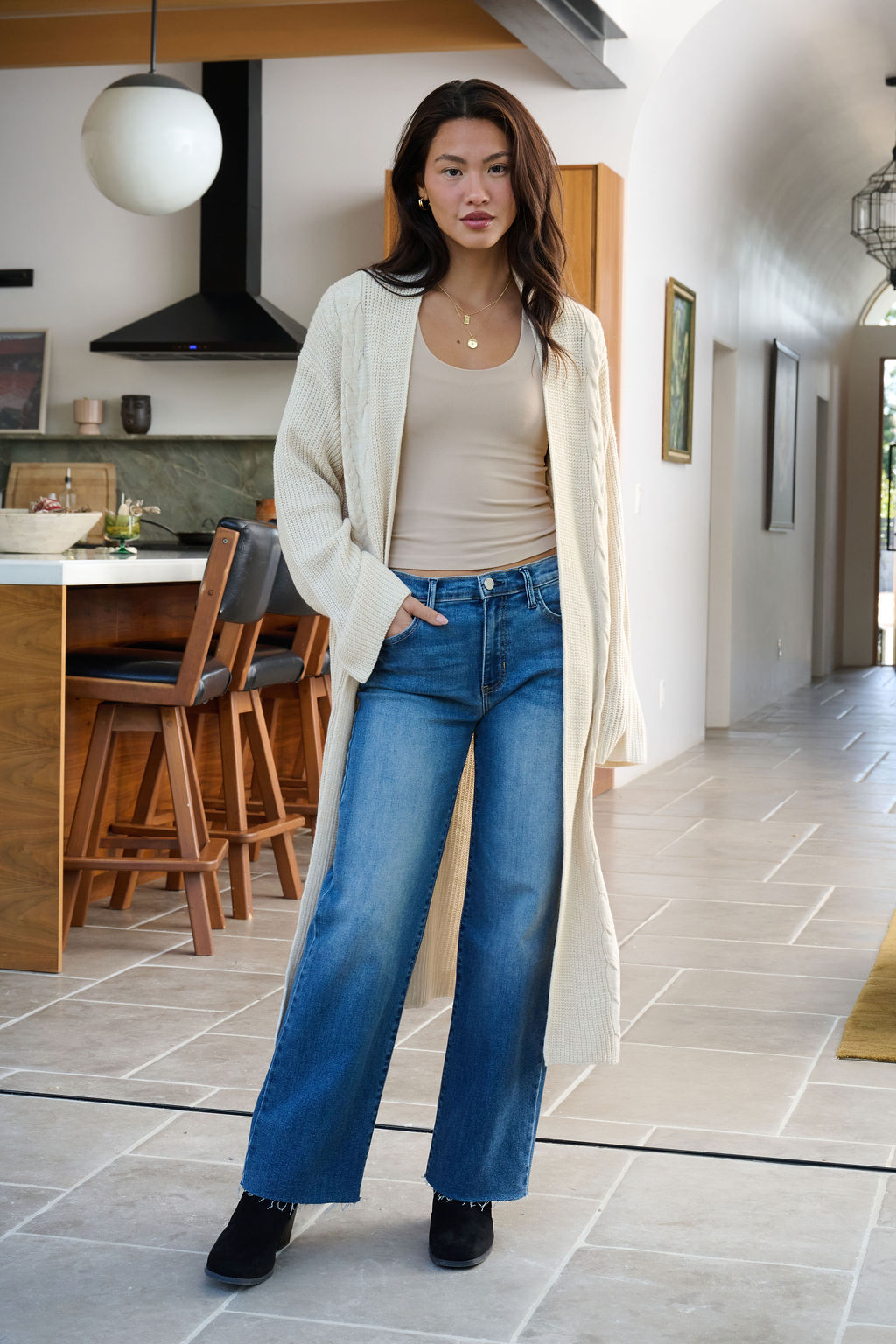 A woman wears the Beige Ribbed Open Front Duster Cardigan indoors with a tan top, blue jeans, and black shoes—ideal for winter layering.