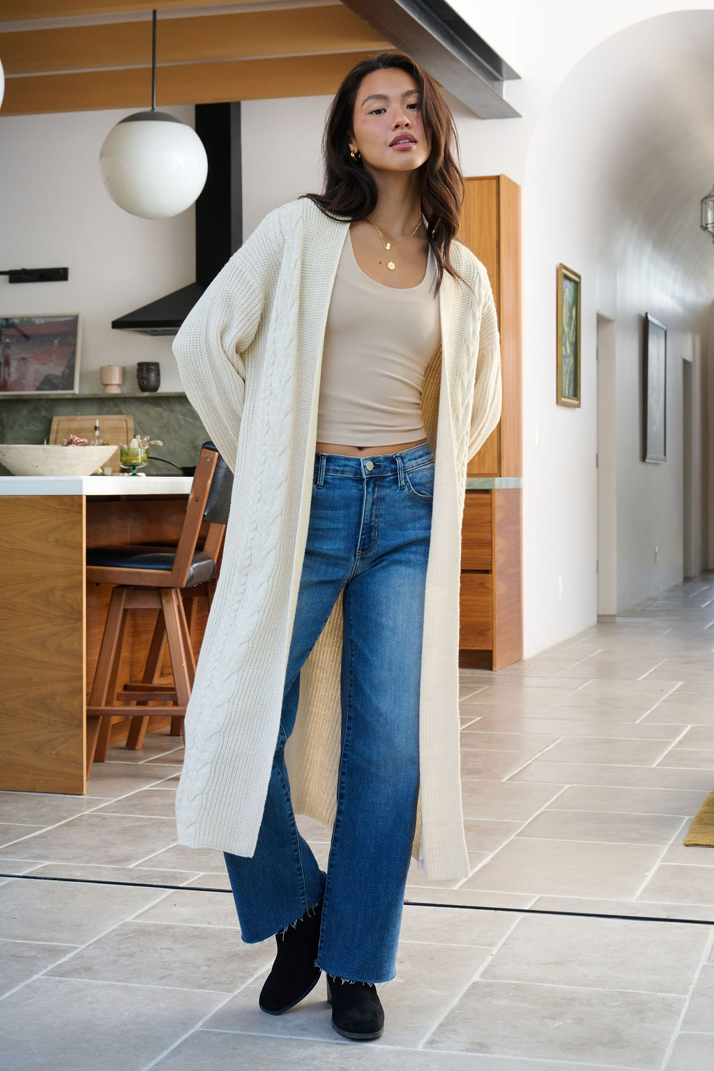 A woman in a Beige Ribbed Open Front Duster Cardigan, blue jeans, and black boots stands in a modern neutral-toned kitchen.