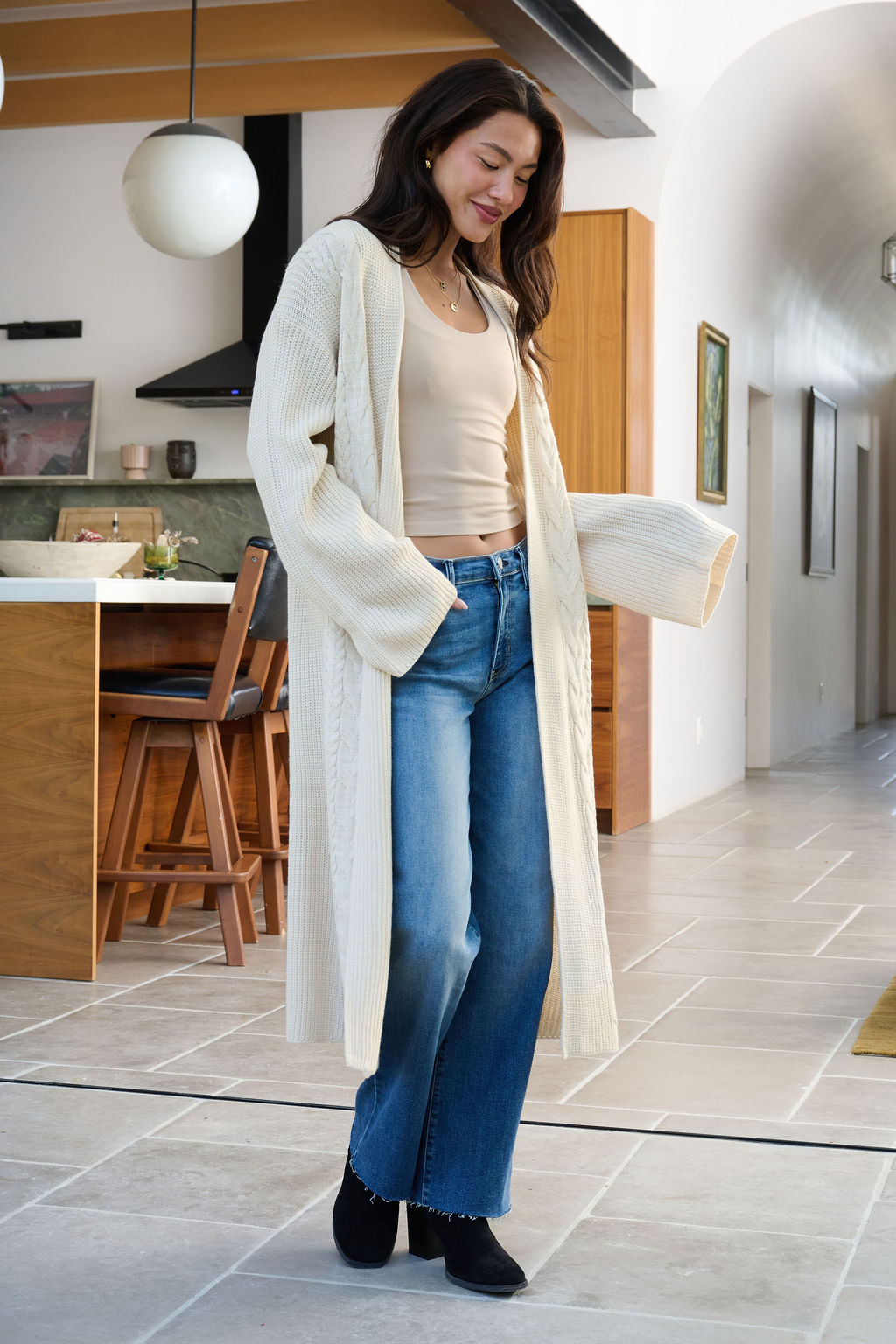A woman smiles while winter layering in a modern kitchen, wearing a Beige Ribbed Open Front Duster Cardigan, jeans, and black boots.
