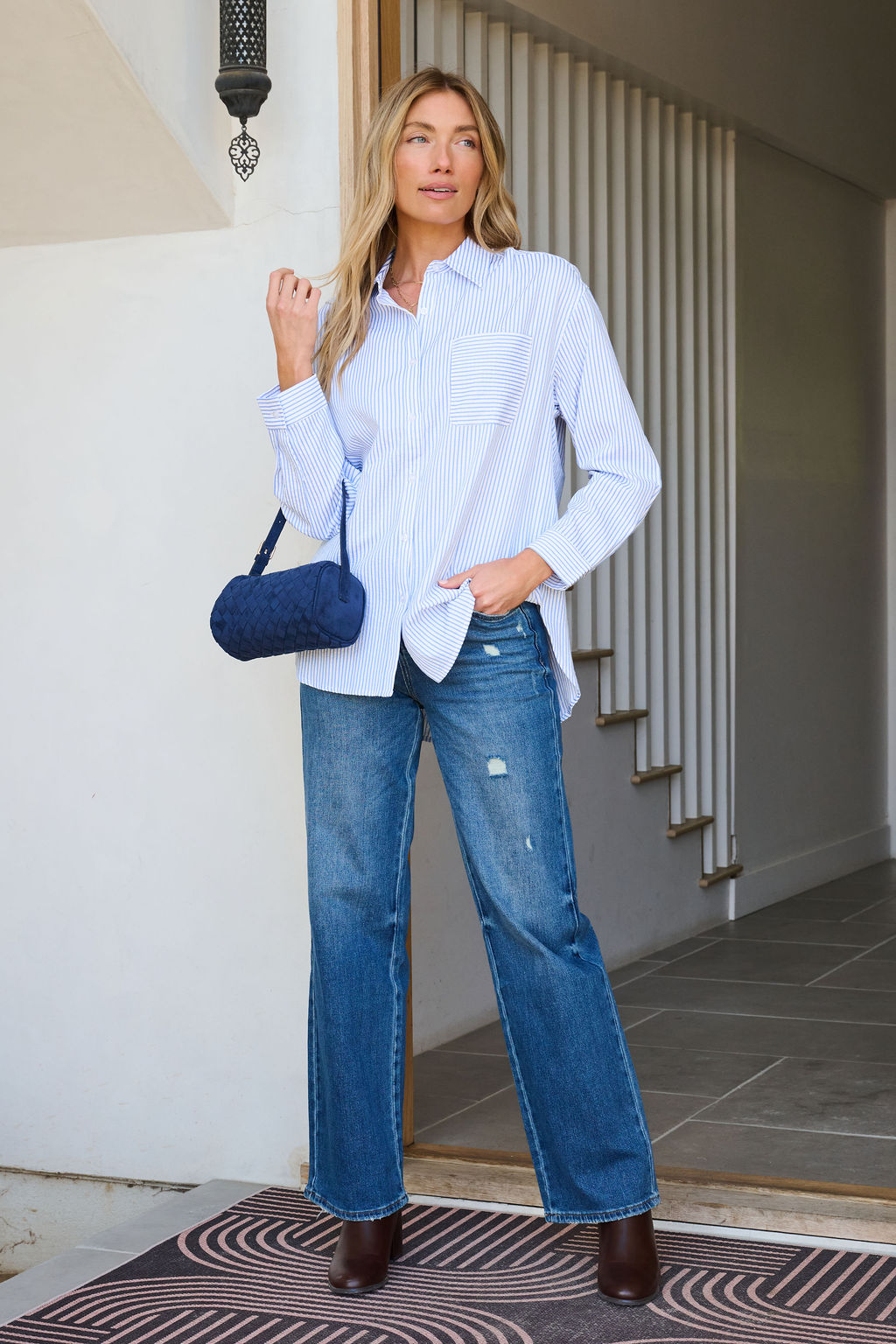 Woman stands in a doorway wearing the Blue and White Striped Open Back Tie Top, blue jeans, brown boots, and holding a blue purse.