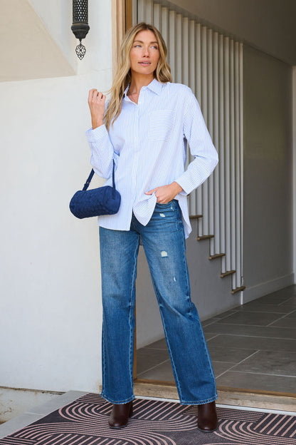 Woman stands in a doorway wearing the Blue and White Striped Open Back Tie Top, blue jeans, brown boots, and holding a blue purse.