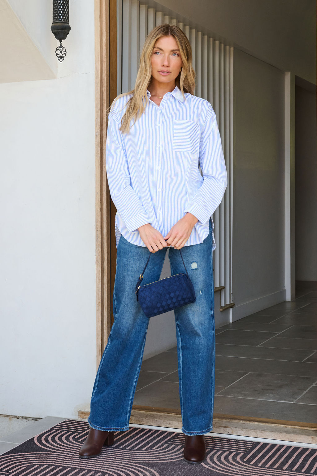 A woman wears the Blue and White Striped Open Back Tie Top indoors, holding a navy clutch and wearing brown boots.