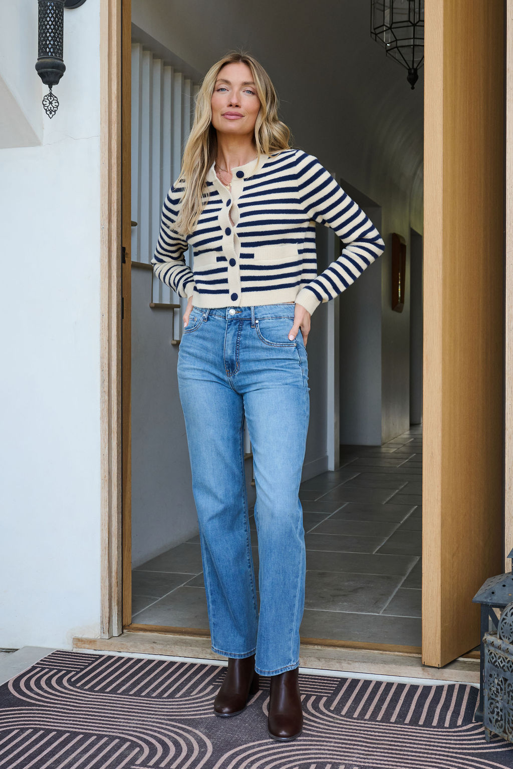 A woman wears a Navy and Tan Striped Button Down Cardigan and blue jeans, standing in a doorway with hands in her pockets, smiling.