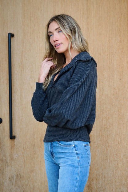 A woman with long blonde hair wears the Ultra Soft Charcoal Collared Pullover and light blue jeans, standing by a wooden wall.