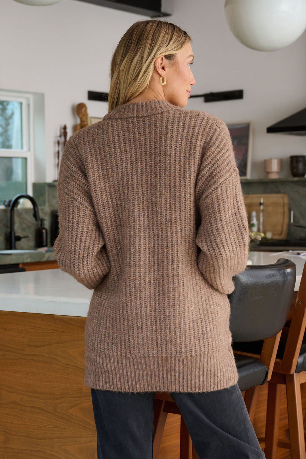 A woman with blonde hair wears a Mocha Heavy Knit Button Down Cardigan in a modern kitchen, facing away from the camera.