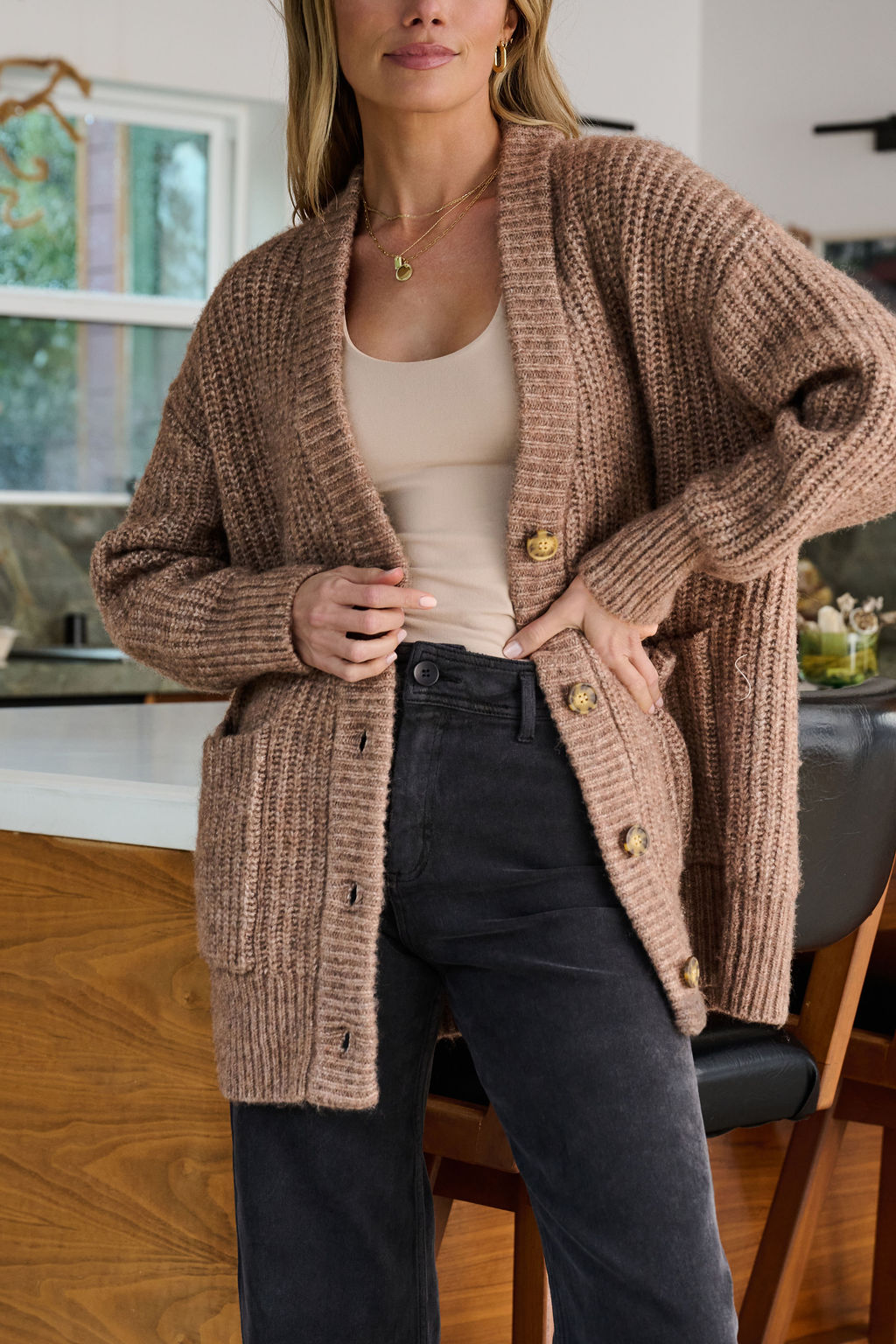 Woman in a Mocha Heavy Knit Button Down Cardigan stands in a modern kitchen, one hand on her hip.