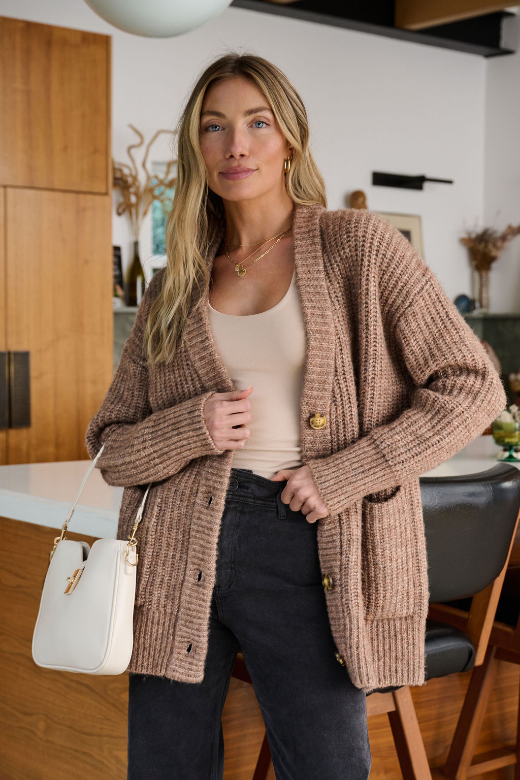 A woman in a beige top, Mocha Heavy Knit Button Down Cardigan, and dark pants stands in a modern kitchen holding a white handbag.
