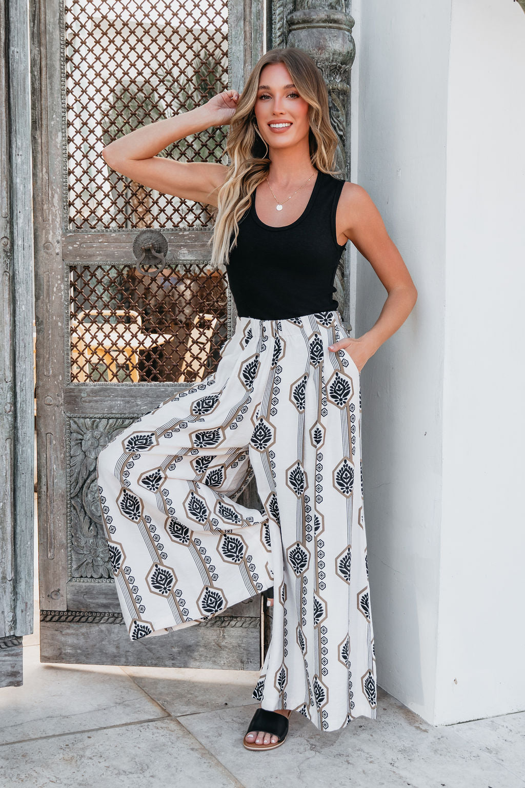Woman in a black tank top and Ivory Print Wide Leg Pants smiles by a decorative gate, one hand on her head.
