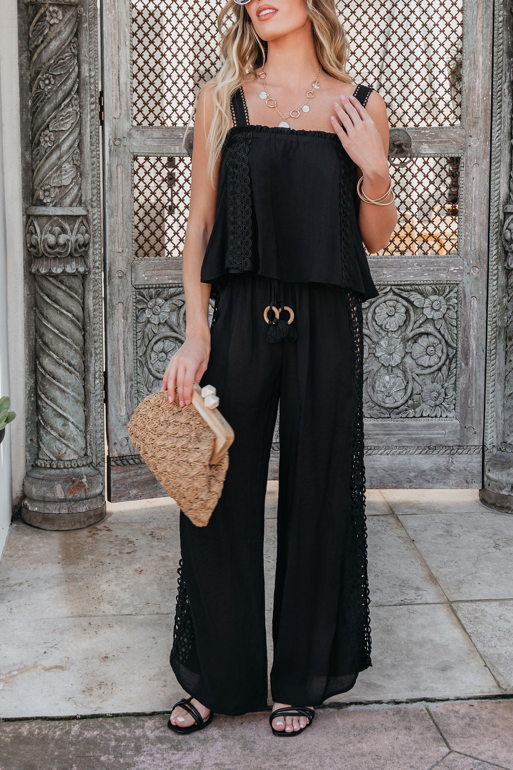 Woman in ELAN Black Lace Trim Wide Leg Pants, holding a woven clutch and wearing sandals, stands before ornate carved doors.