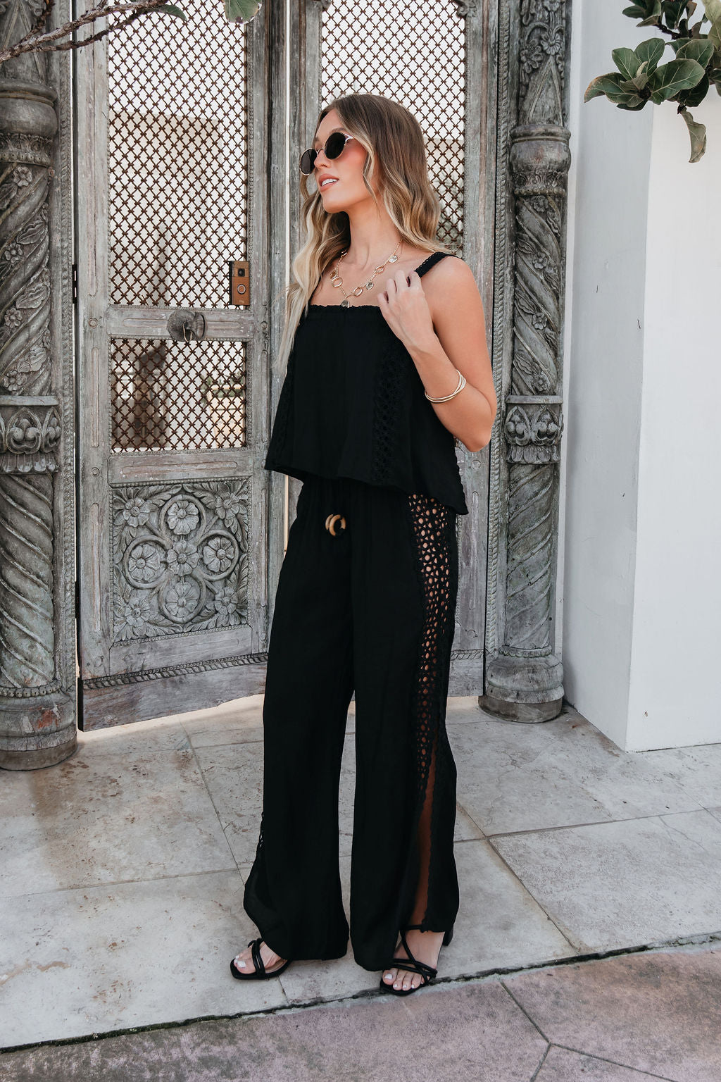 Woman in ELAN Black Lace Trim Wide Leg Pants, sunglasses, and sandals stands by ornate wooden doors outdoors.