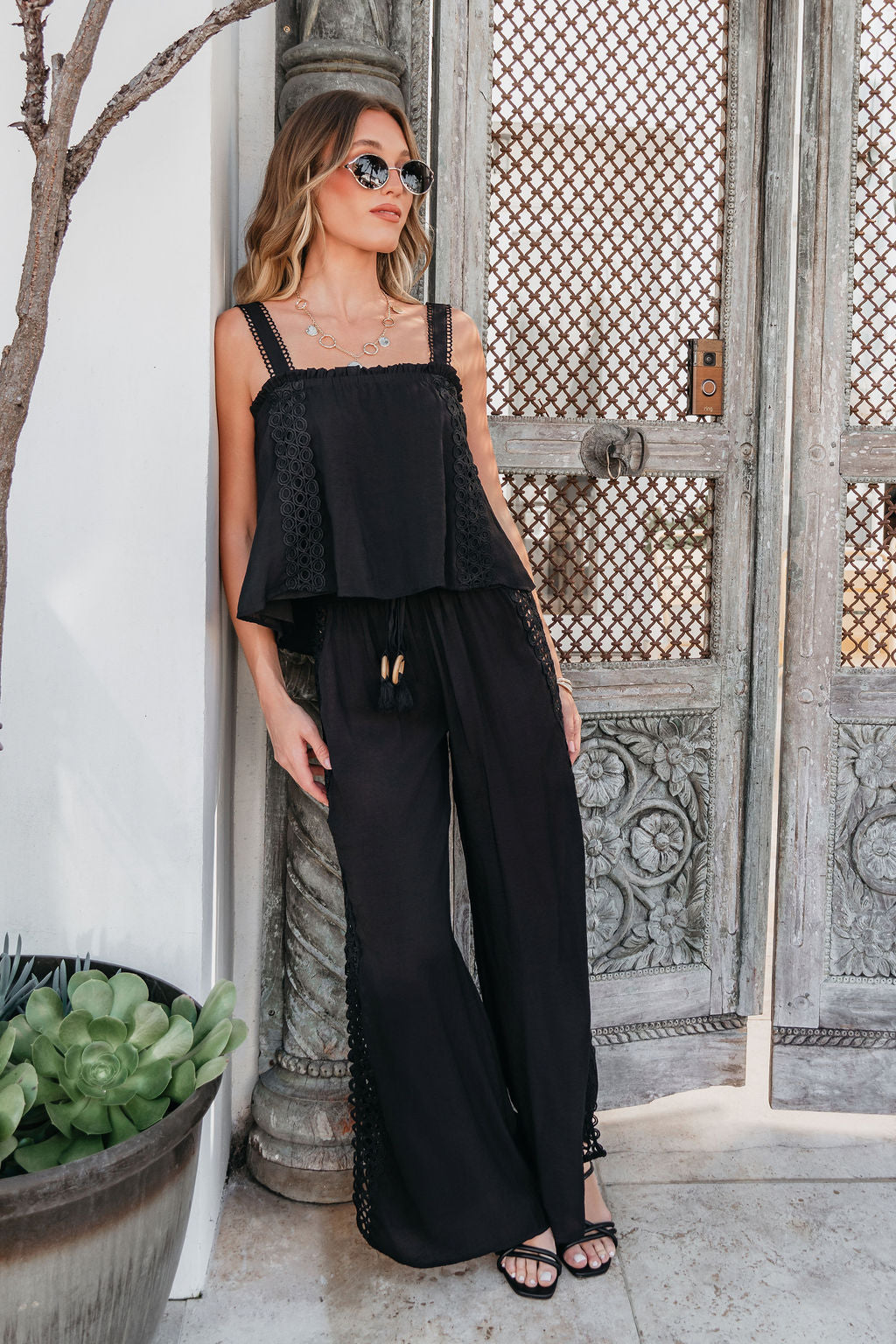 Woman in sunglasses wears ELAN Black Lace Trim Wide Leg Pants, standing by ornate doors next to a large potted plant.