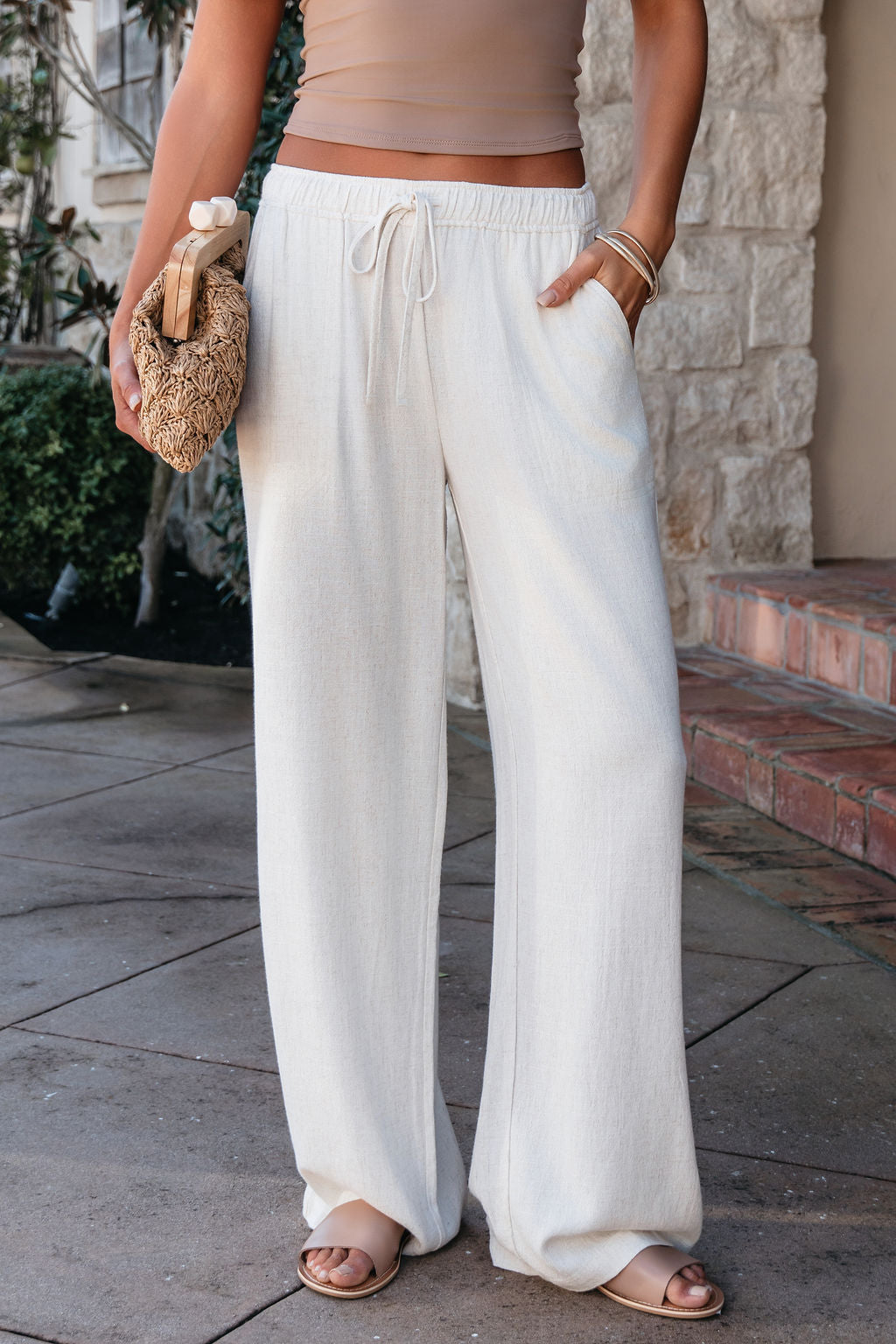 A woman in Natural Drawstring Wide Leg Linen Pants, tan sandals, and a crop top holds a woven clutch on a stone walkway.