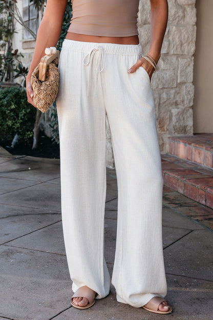 A woman in Natural Drawstring Wide Leg Linen Pants, tan sandals, and a crop top holds a woven clutch on a stone walkway.