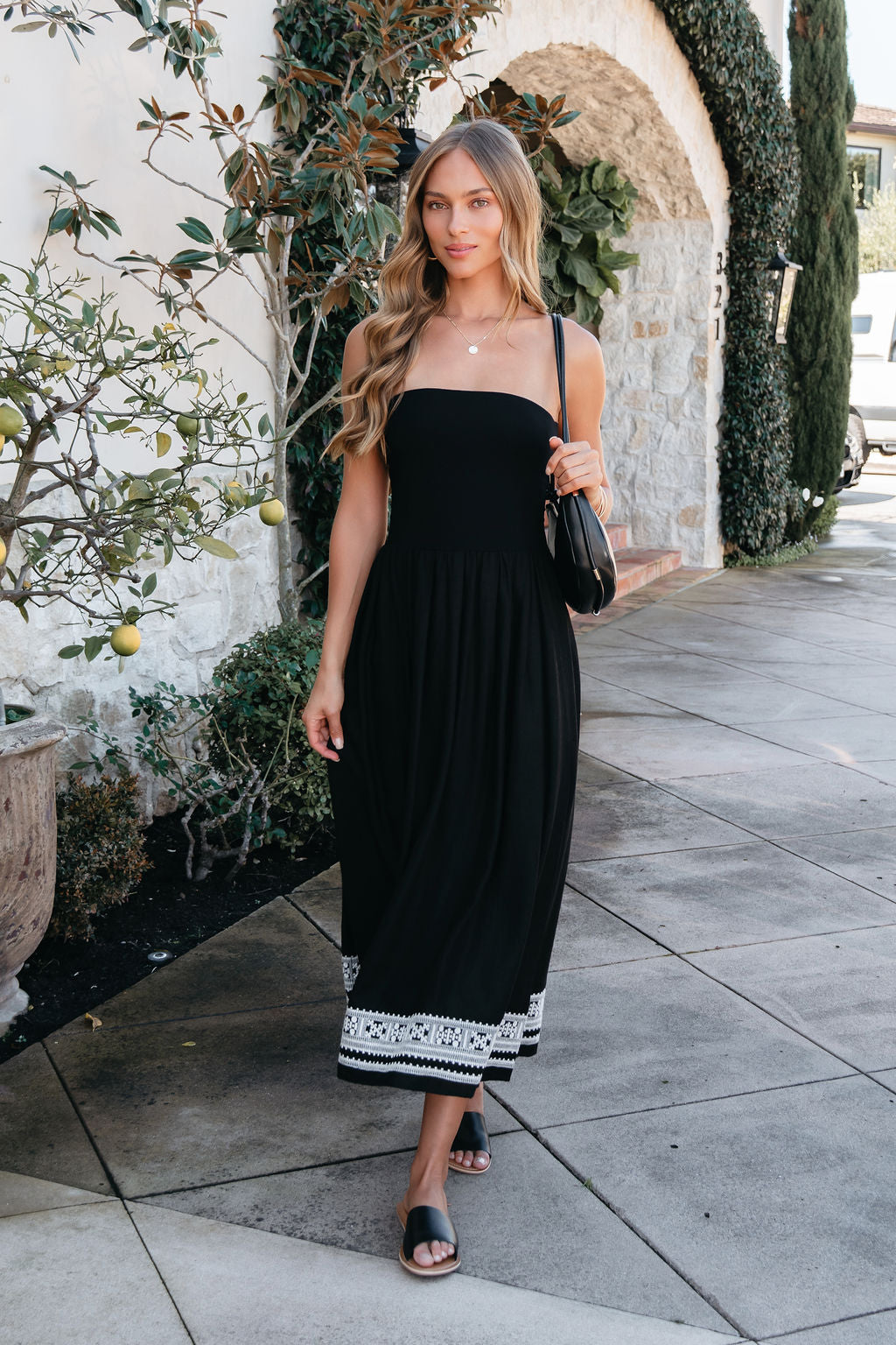 A woman in a Black Strapless Embroidered Midi Dress walks on a sidewalk near a stone building and potted plant.