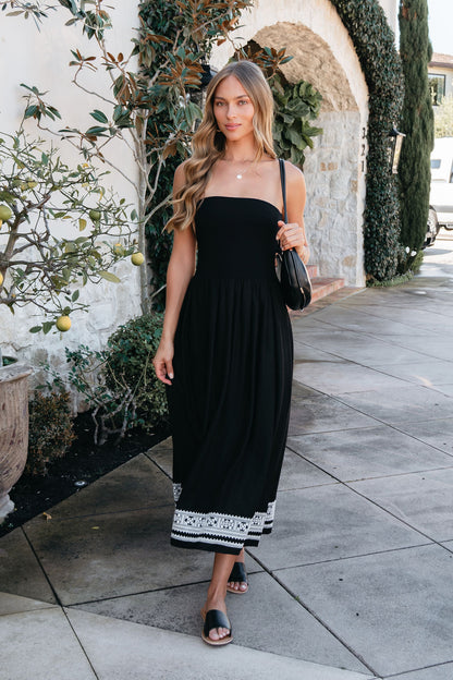A woman in a Black Strapless Embroidered Midi Dress walks on a sidewalk near a stone building and potted plant.