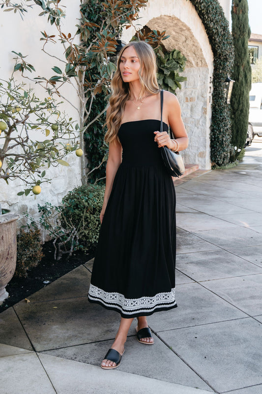 Woman in a Black Strapless Embroidered Midi Dress walks in sandals with a bag along a stone path next to greenery.