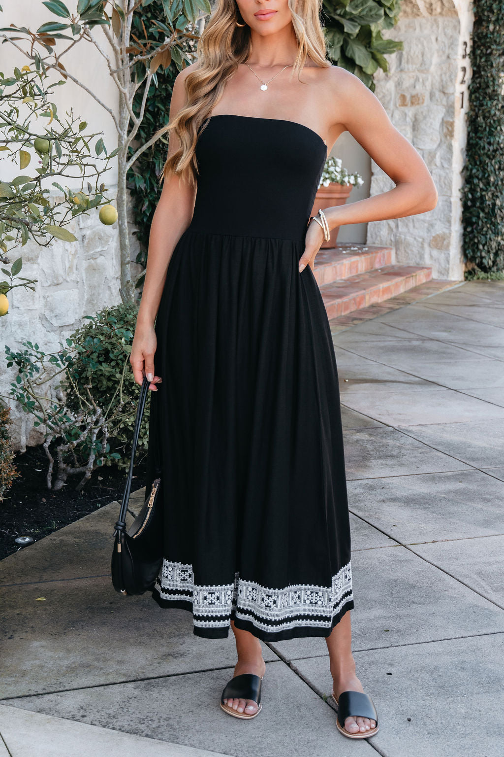 Woman in a Black Strapless Embroidered Midi Dress, black sandals, and a black bag stands on a stone walkway.