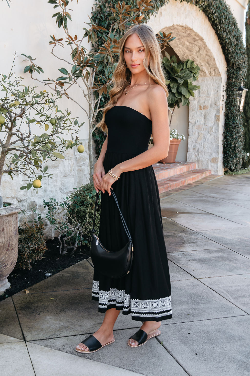 A woman wears the Black Strapless Embroidered Midi Dress outside, holding a black purse and wearing black sandals.