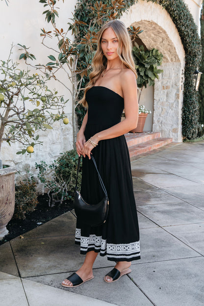 A woman wears the Black Strapless Embroidered Midi Dress outside, holding a black purse and wearing black sandals.