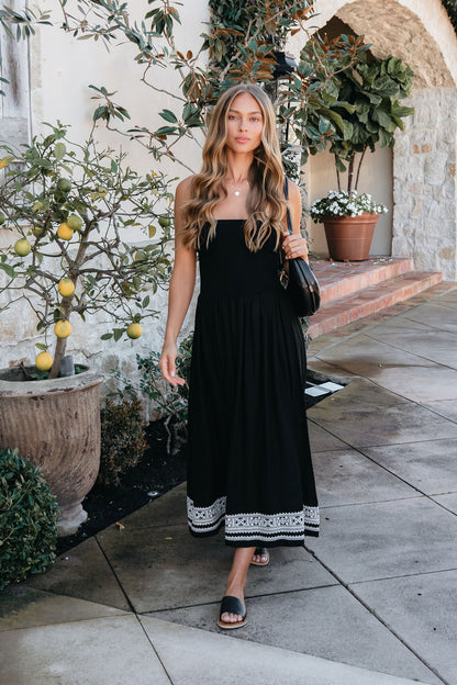 Woman with long wavy hair wears the Black Strapless Embroidered Midi Dress and sandals, walking by potted plants and a stone wall.