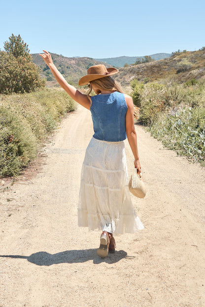 A woman in the Weston Button Front Denim Vest, white skirt, and hat strolls a rural path with a woven bag, raising one arm.