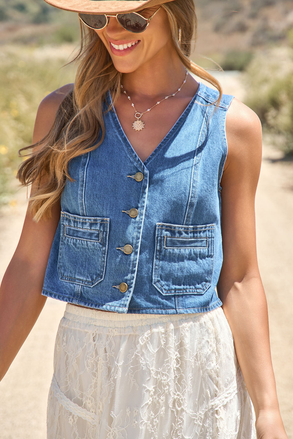 Woman in sunglasses and a hat wears the Weston Button Front Denim Vest with a white lace skirt outdoors on a sunny day.