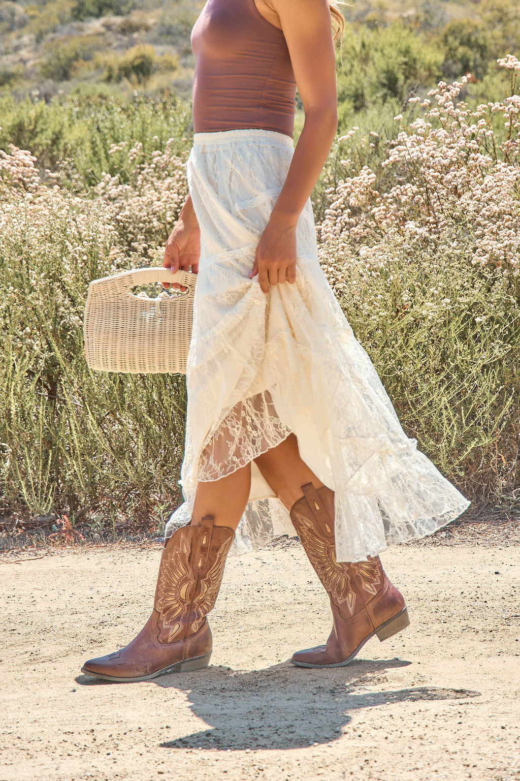 Wearing the Rio Lace Tiered Midi Skirt in cream, a person walks outdoors with a brown tank and woven handbag.