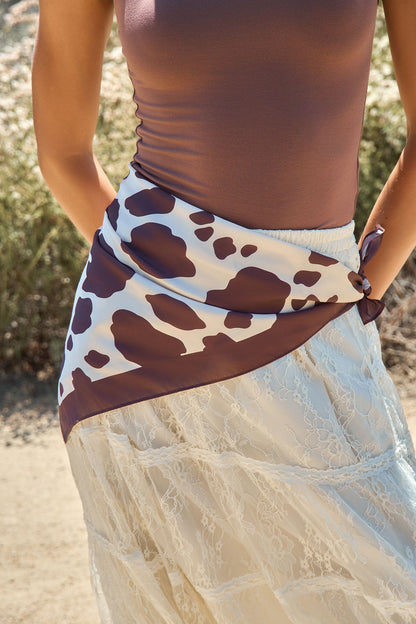 A person wears a mauve top, lace skirt, and the Cow Print Bandana Scarf - Brown tied at the waist, standing outdoors.