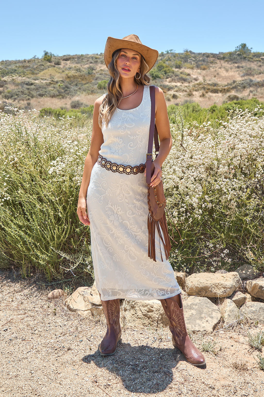 Woman in the Tucson Floral Lace Midi Dress - Ivory stands by wildflowers on a sunny day, wearing a cowboy hat and brown boots.