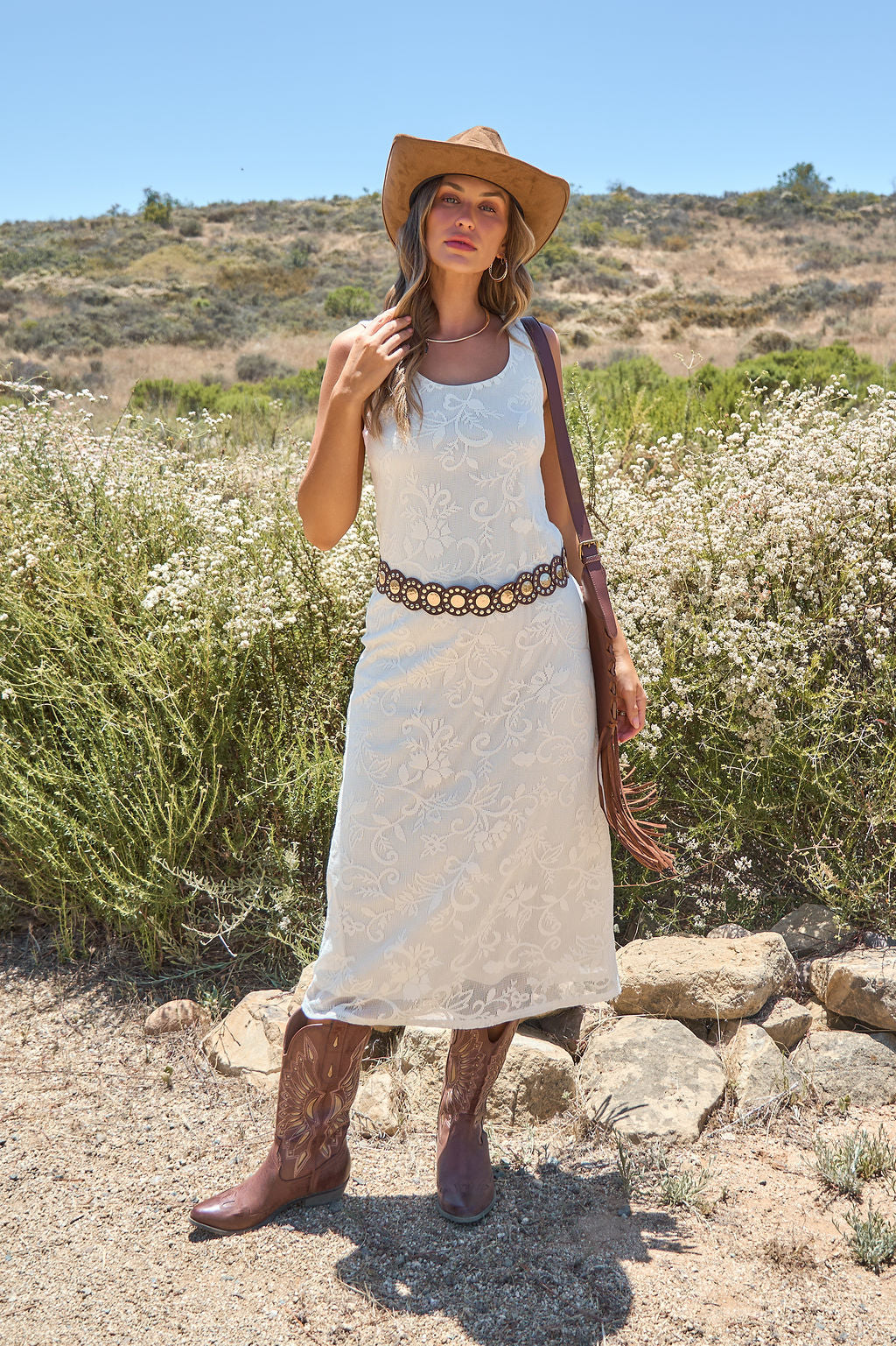 Woman in the Tucson Floral Lace Midi Dress - Ivory, cowboy boots, and hat stands outdoors among greenery on a sunny day.