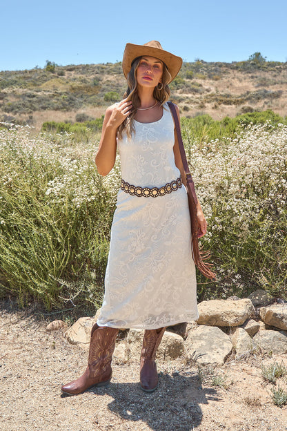 Woman in the Tucson Floral Lace Midi Dress - Ivory, cowboy boots, and hat stands outdoors among greenery on a sunny day.