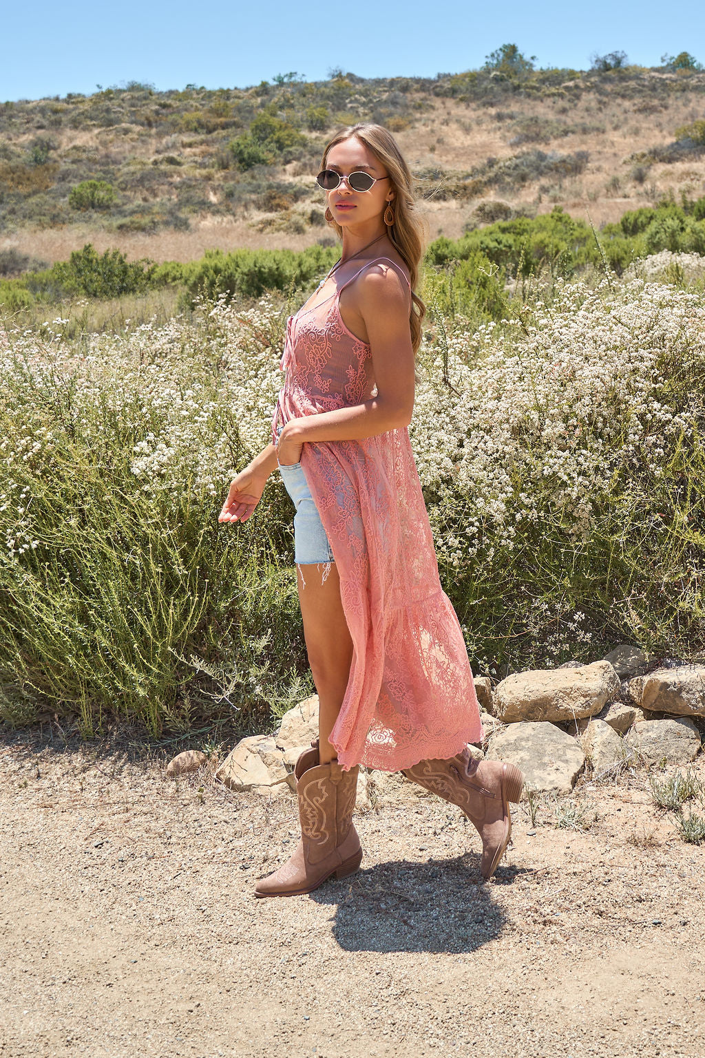 Woman in sunglasses and denim walks a dirt path, layering her Desert Rose Lace Midi Vest amid green festival hills.