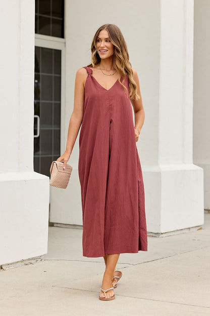 Woman in burgundy Audrey Knot Strap Midi Dress, sandals, wavy hair, and small handbag stands by a white building on the sidewalk.