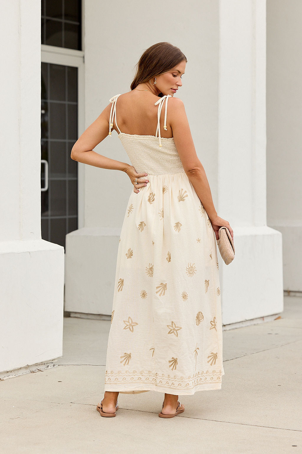Woman in the Natural Beauty Embroidered Maxi Dress, cream with gold patterns and tie straps, holds a clutch near a white building.