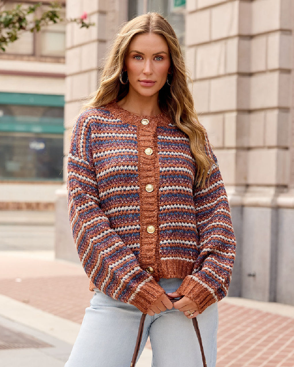 A woman wearing a chunky knit cardigan with horizontal stripes in rust orange, navy blue, and cream, featuring gold-tone buttons down the front. She is paired with light-wash jeans, standing outdoors in front of a stone building.