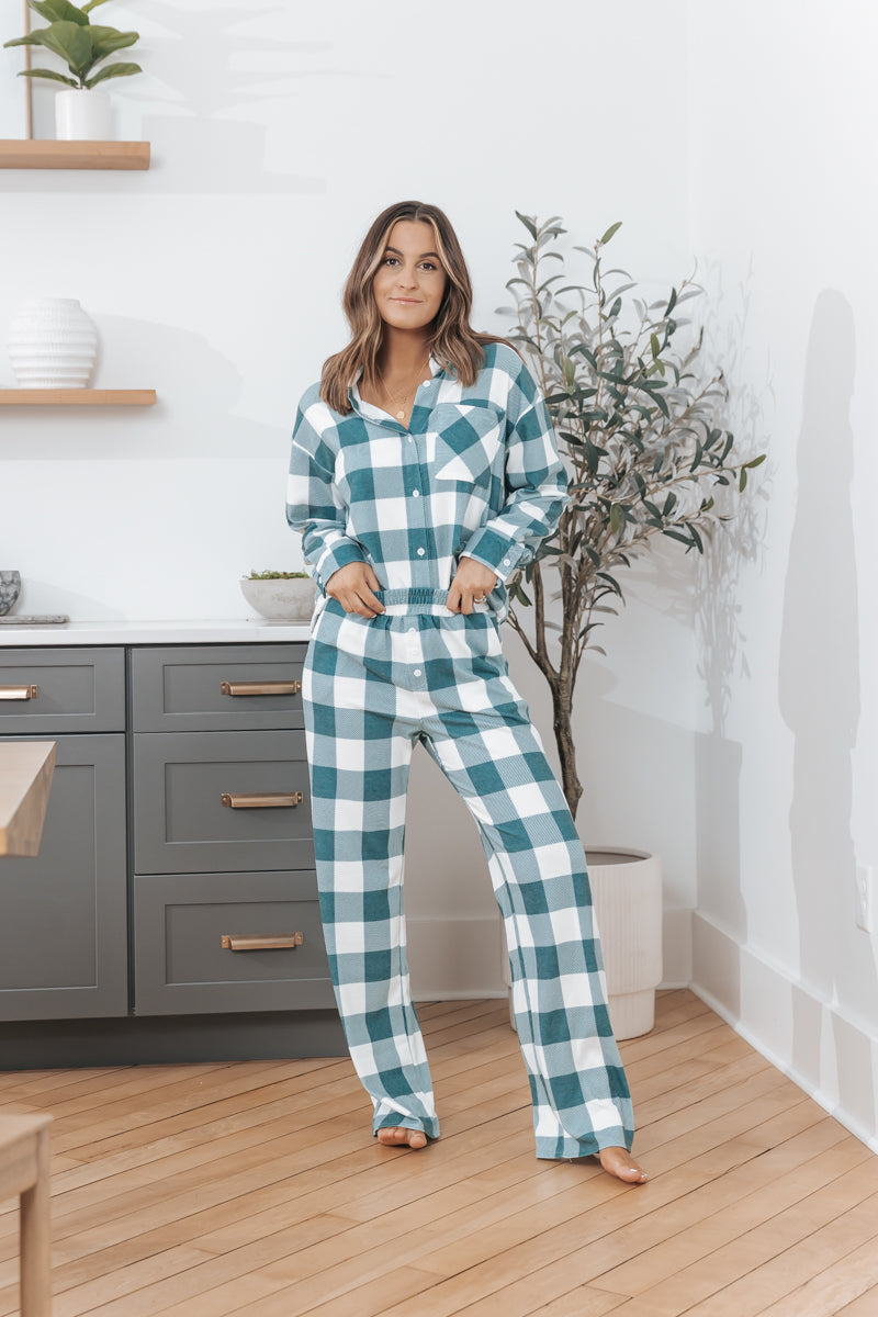 Woman in kitchen wears the Rest Easy Green Plaid PJ Set, hands in pockets, standing by a potted plant.