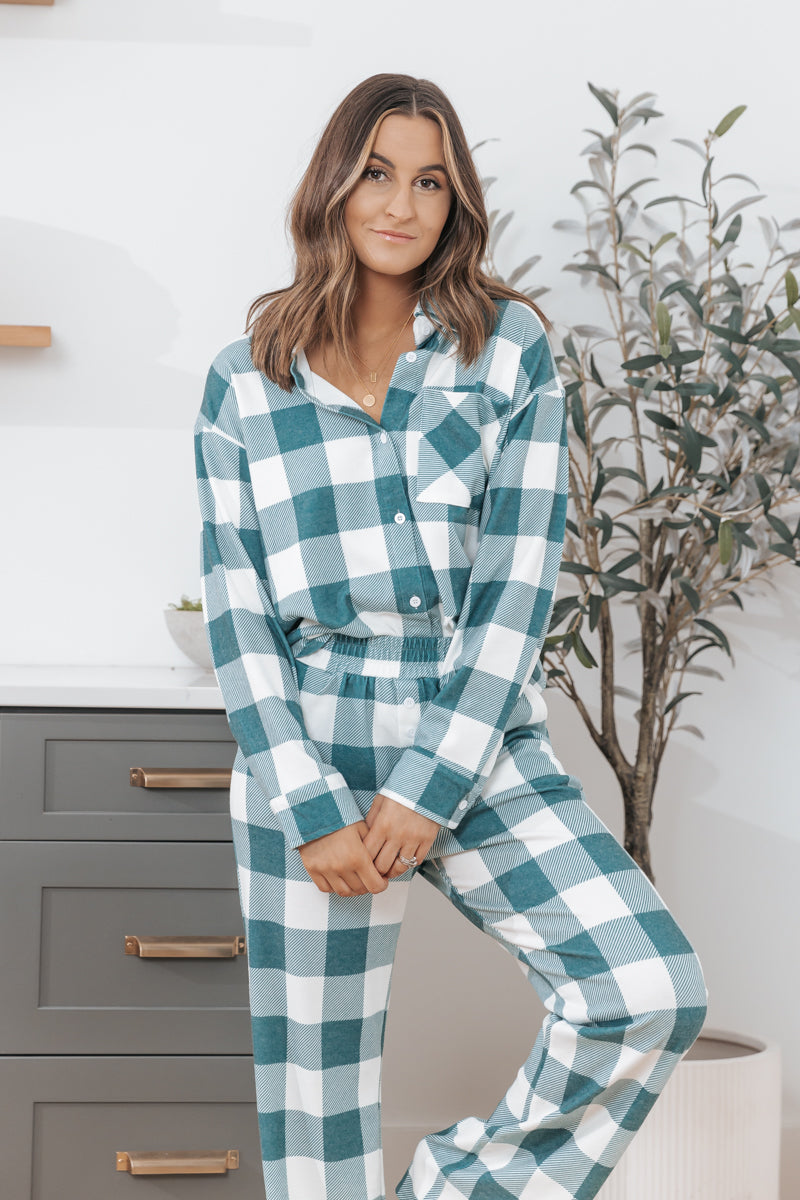 A woman wears the Rest Easy Green Plaid PJ Set indoors, leaning on a counter with a potted plant in the background.
