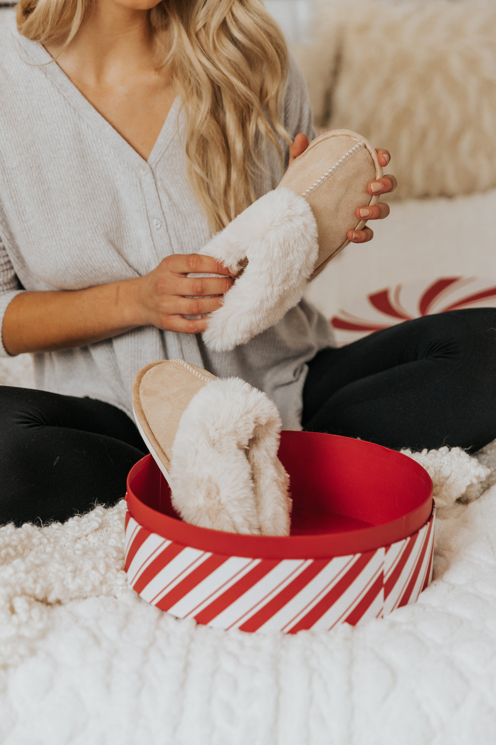 A woman holds Sweet Dreams Sandy Faux Sheepskin Slippers - FINAL SALE next to a red and white gift box on a cozy bed.