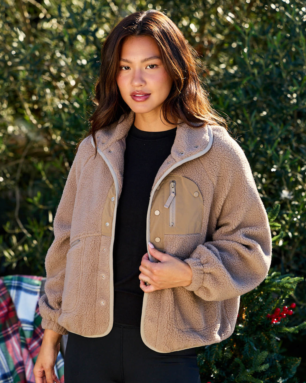 Woman wearing a beige fleece jacket with a black shirt underneath, standing outdoors with greenery in the background.