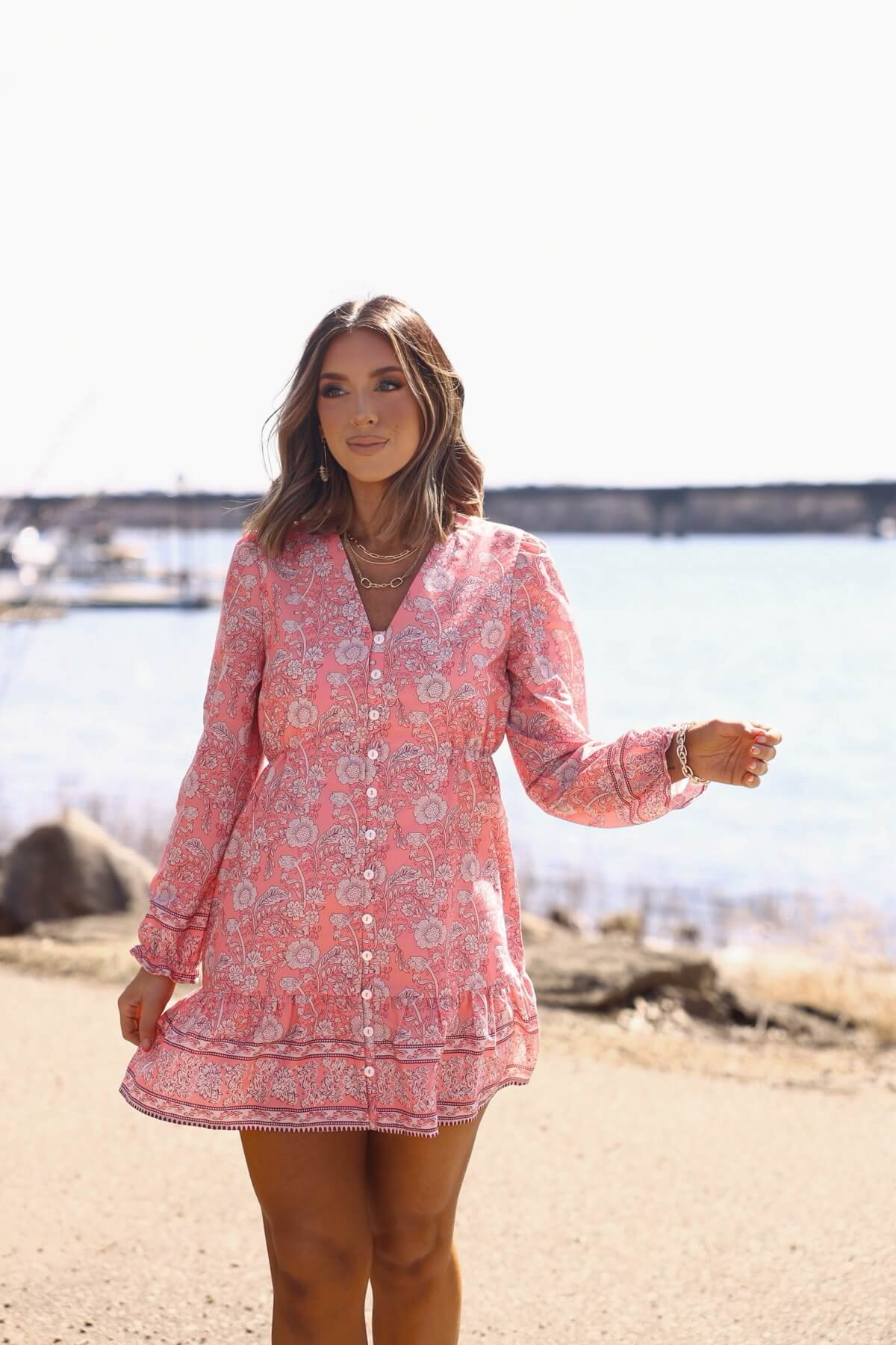 A woman in a Rosie Pink Floral Print Buttoned Mini Dress stands by the water, boats and rocky shore in the background.