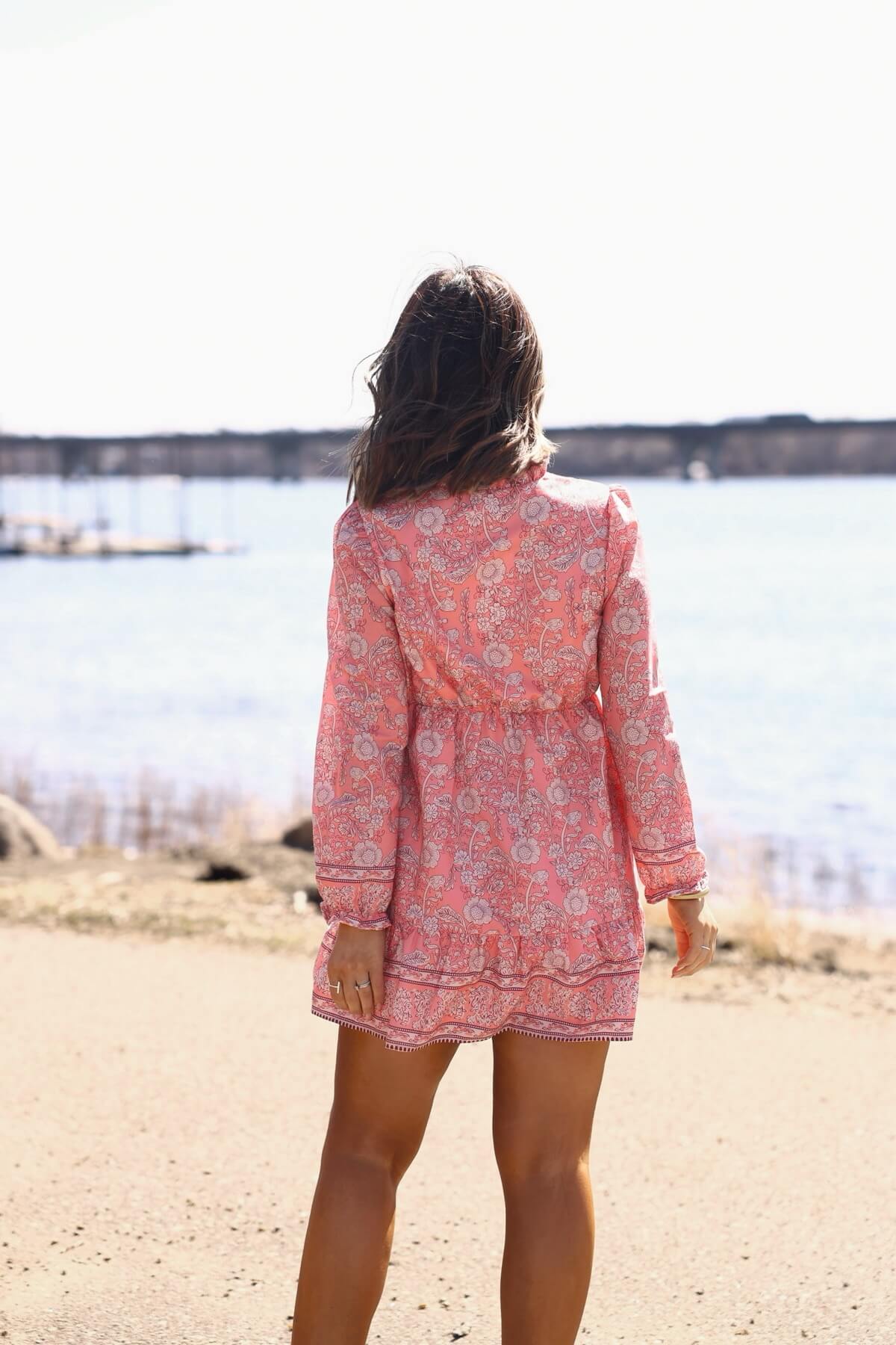 Woman in a Rosie Pink Floral Print Buttoned Mini Dress stands by a lake, her back to the camera on a sunny day.