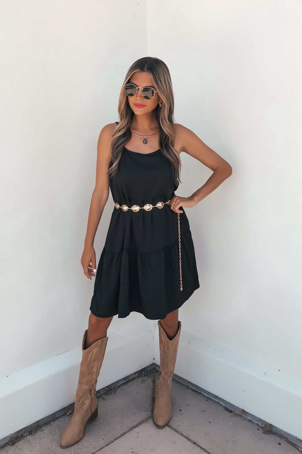Woman in the On Point Tiered Mini Dress - Black, gold belt, tan knee-high boots, sunglasses, and red lipstick against a white wall.