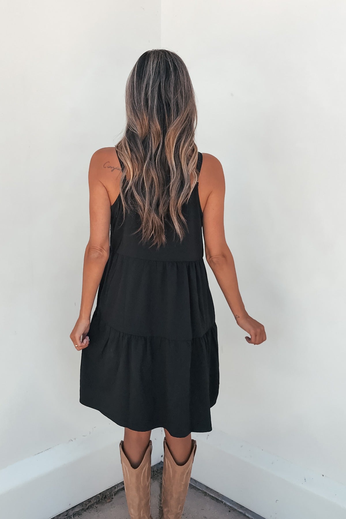 Woman with long wavy hair wears the On Point Tiered Mini Dress - Black, facing a white wall, back to camera.