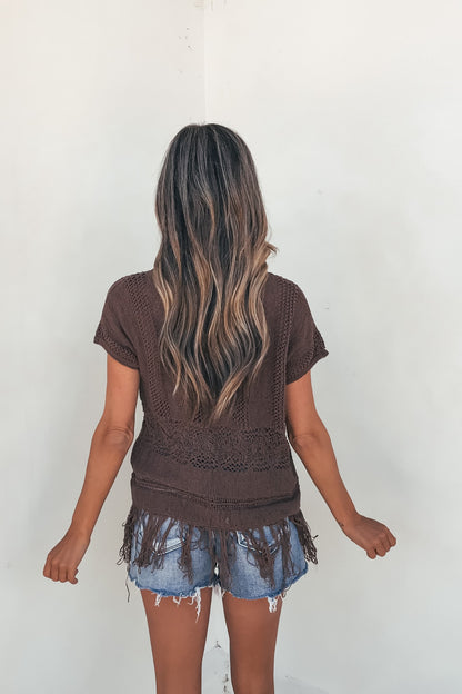 A woman with long wavy hair wears the Boho Fringe Sweater - Brown and denim shorts, standing by a plain light wall.