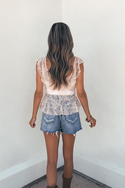 A woman with wavy brown hair wears the Steve Madden Kaesey Ruffle Top in rose and denim shorts, facing a white wall.