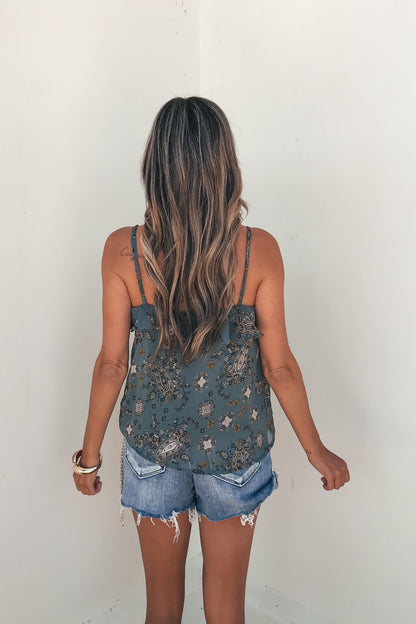 A woman with long wavy hair wears a Teal Paisley Print Ruffled Cami and distressed denim shorts, facing a white wall.