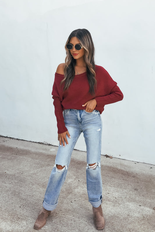 Woman in sunglasses, Off The Shoulder Ribbed Sweater - Wine, ripped jeans, and boots stands on pavement by a white wall.
