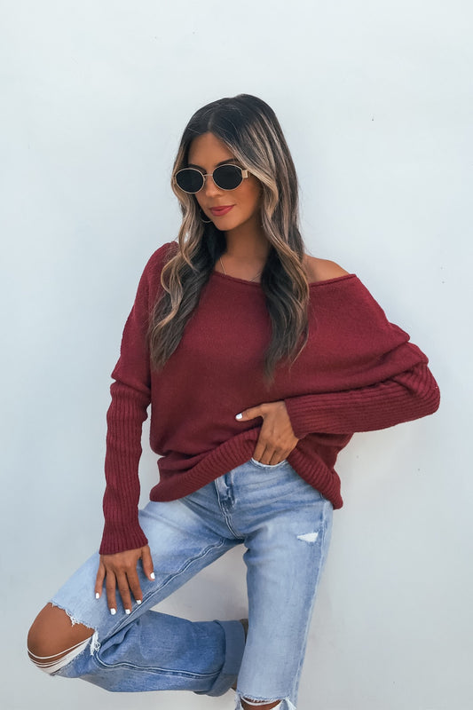 Woman in sunglasses models Off The Shoulder Ribbed Sweater - Wine with ripped jeans against a plain white wall.