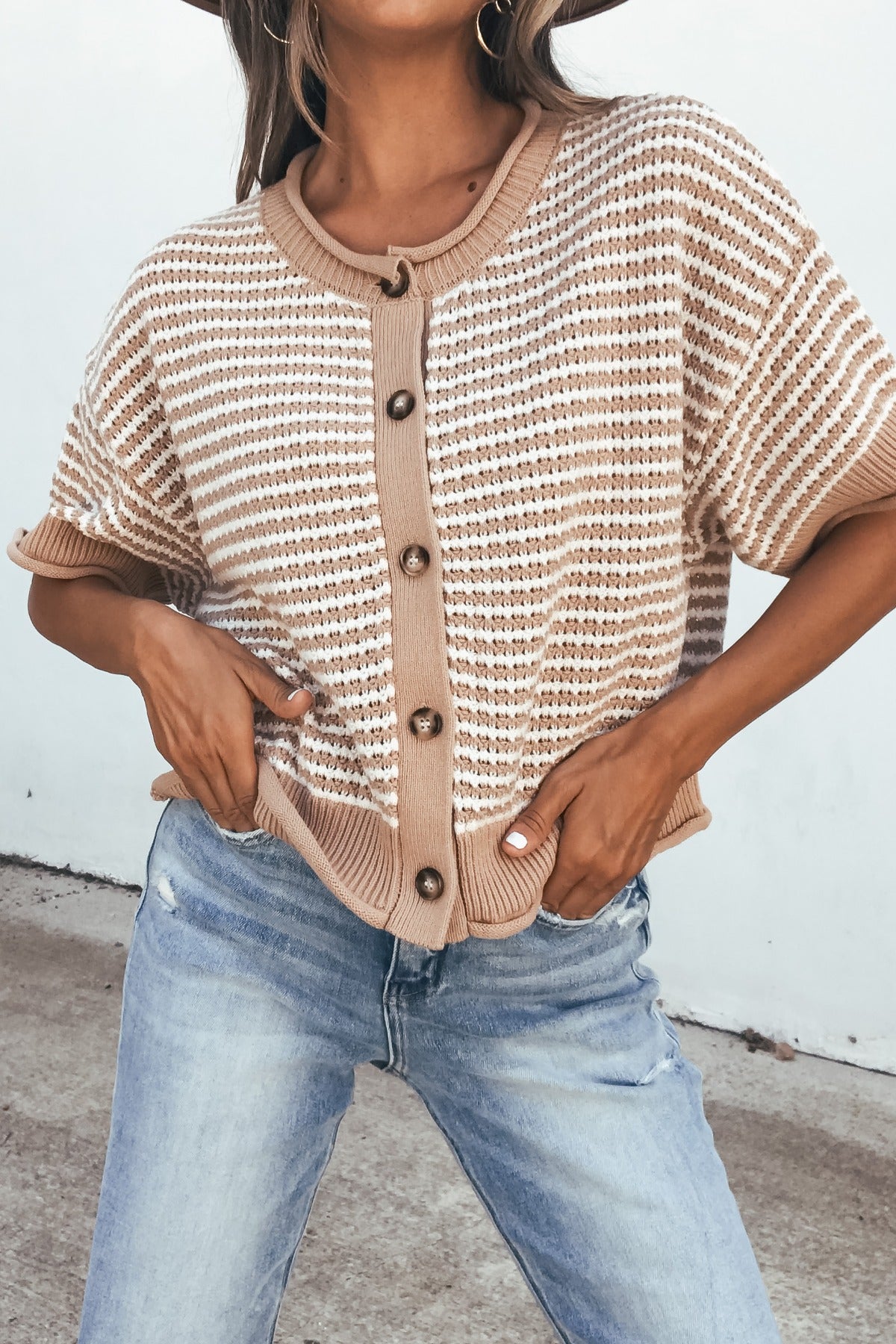 Woman in a tan striped button down sweater and light blue jeans, standing outdoors.