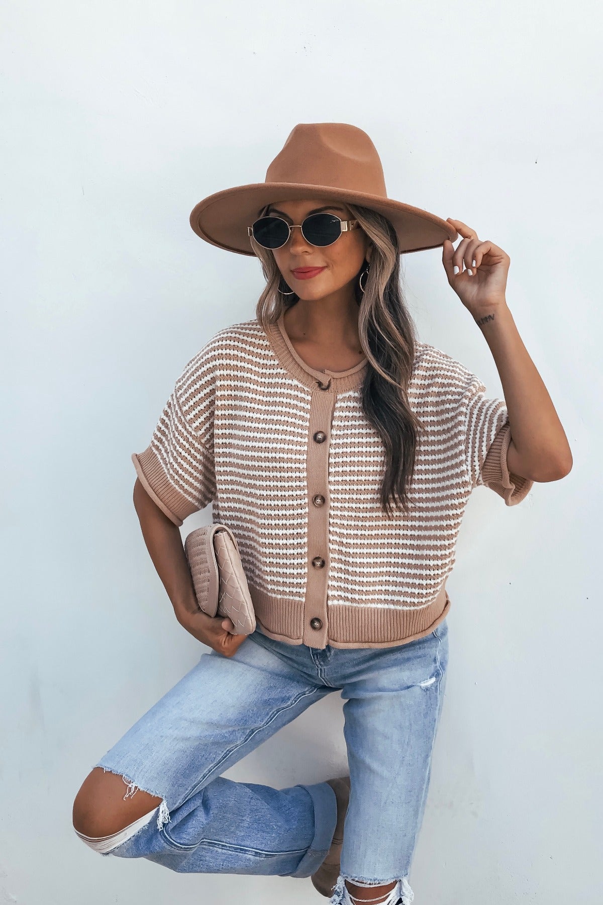 Woman in tan striped button down sweater, hat, and sunglasses poses with clutch and ripped jeans against a white wall.